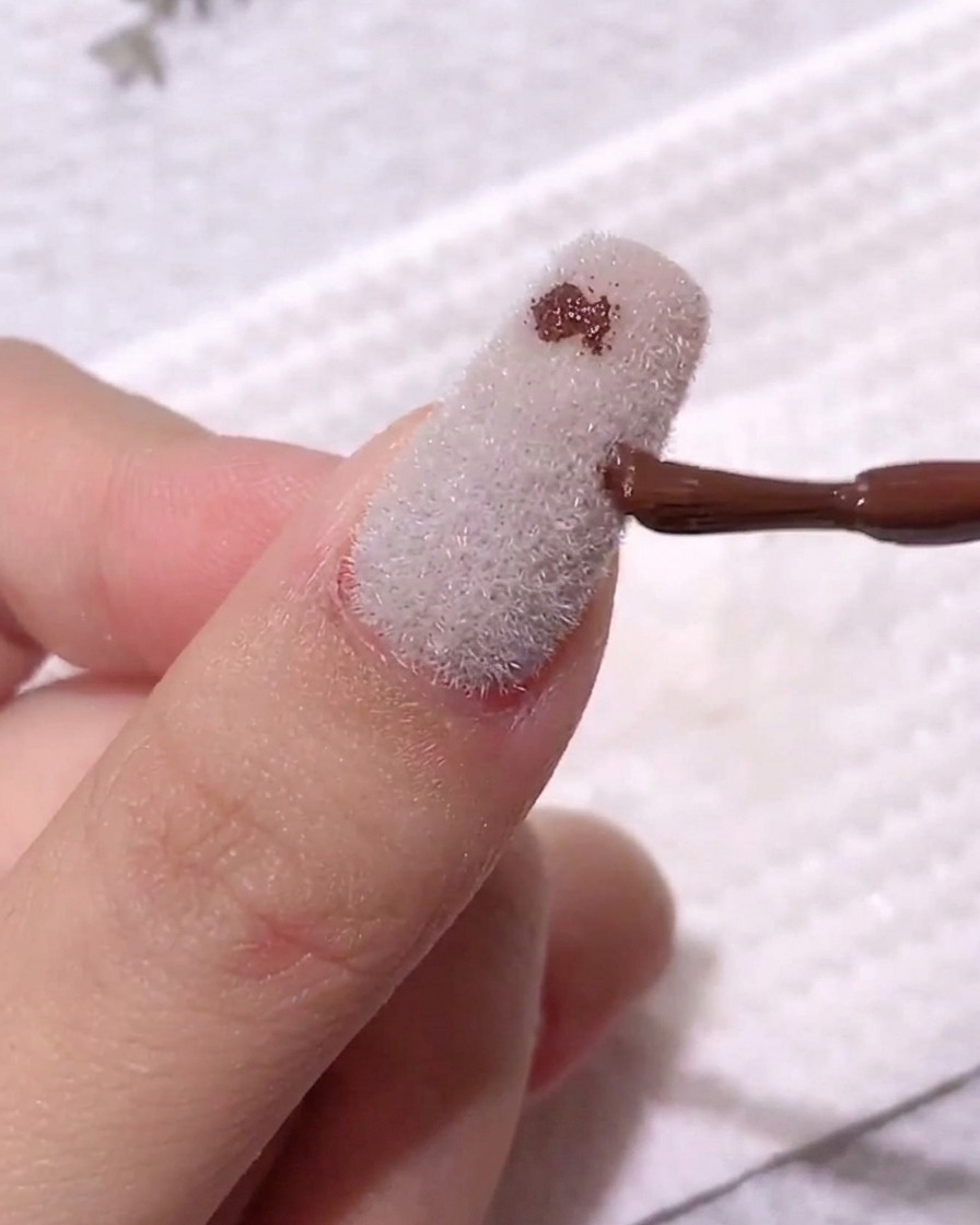 A nail brush depositing a small, dark coffee-colored gel dot onto a matte, white velvet-textured nail surface.