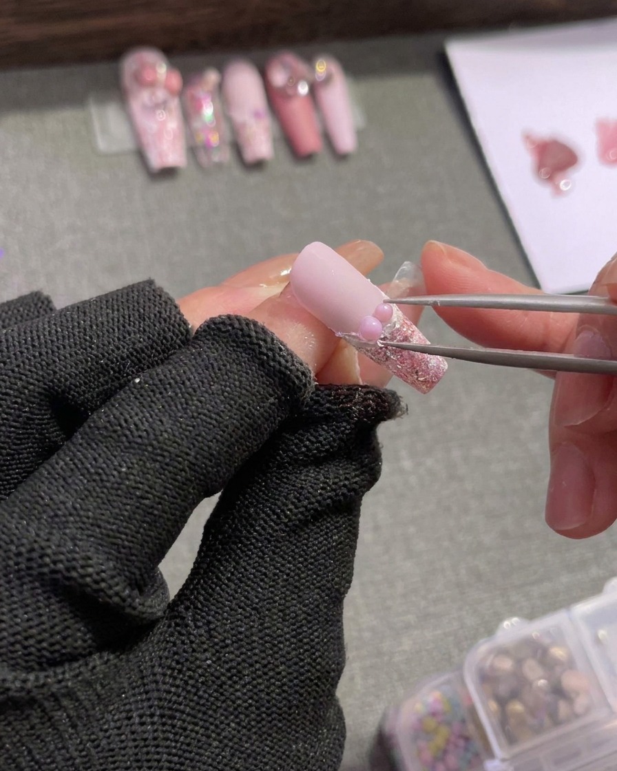 Tweezers carefully positioning tiny silver rhinestones and a small pink bow charm next to a pearl on the French tip border.