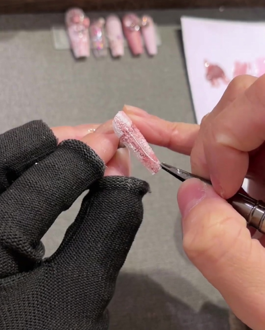 A close-up shot of a long liner brush applying thin white lines over a painted plaid nail design.