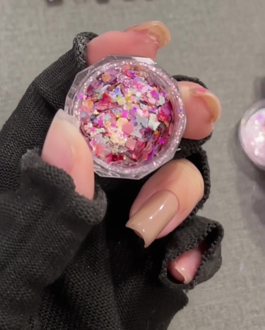 A close-up of a flat brush picking up colorful, chunky sequins from a small round pot.