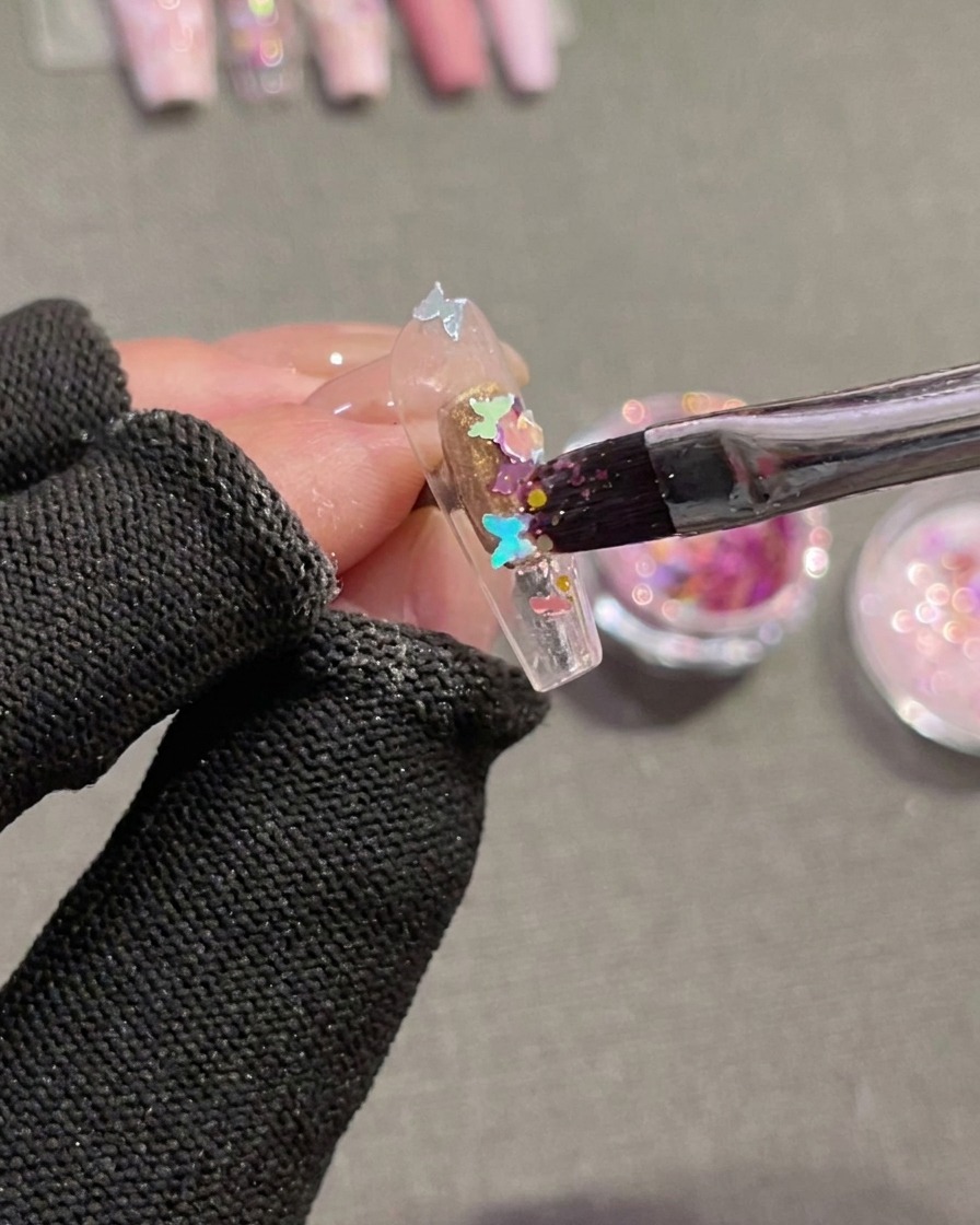 A close-up view of a brush being used to press holographic butterfly-shaped sequins onto a clear nail tip coated in wet, uncured gel.