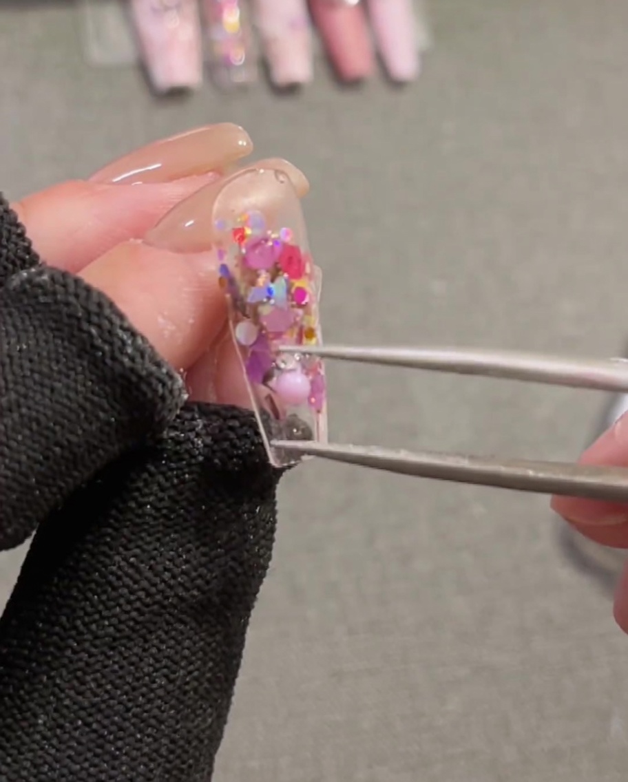 Close-up of tweezers holding and positioning a 3D pink ribbon charm onto a nail that has wet builder gel on it.