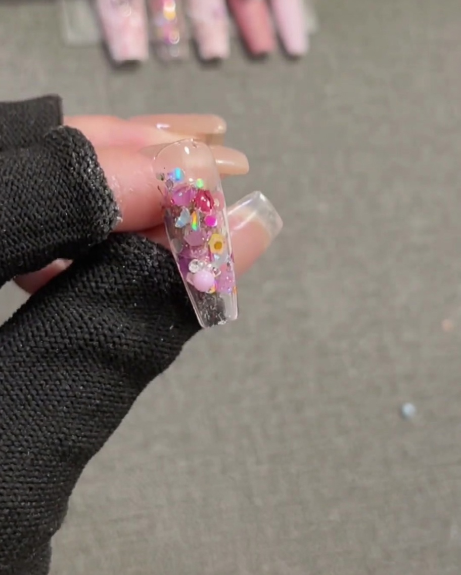 A brush applying a final layer of clear, high-gloss top coat over a completed nail featuring embedded sequins and a 3D pink bow charm.