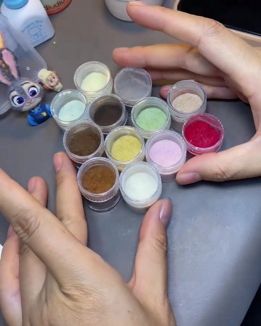 A hand holding tweezers selecting a small clear container filled with light yellow velvet flocking powder.