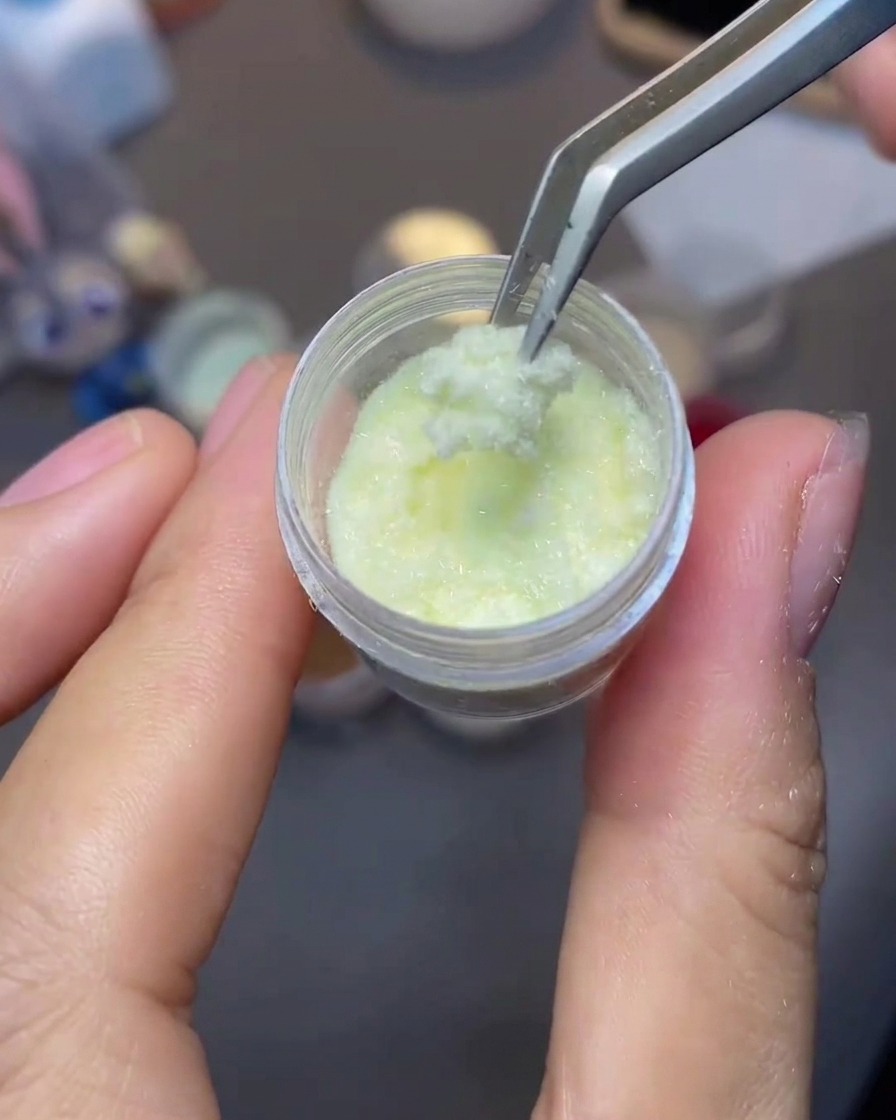 Close-up of tweezers picking up a pinch of light yellow velvet powder from an open jar.