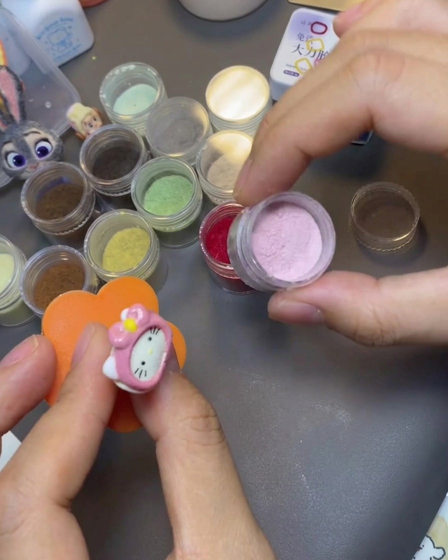 A hand dipping a Hello Kitty nail charm into a jar of pink velvet powder.