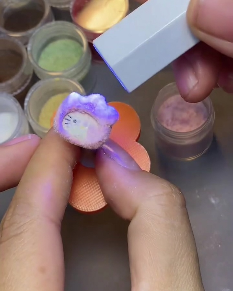 A hand tapping a cured Hello Kitty nail charm to remove excess velvet powder.