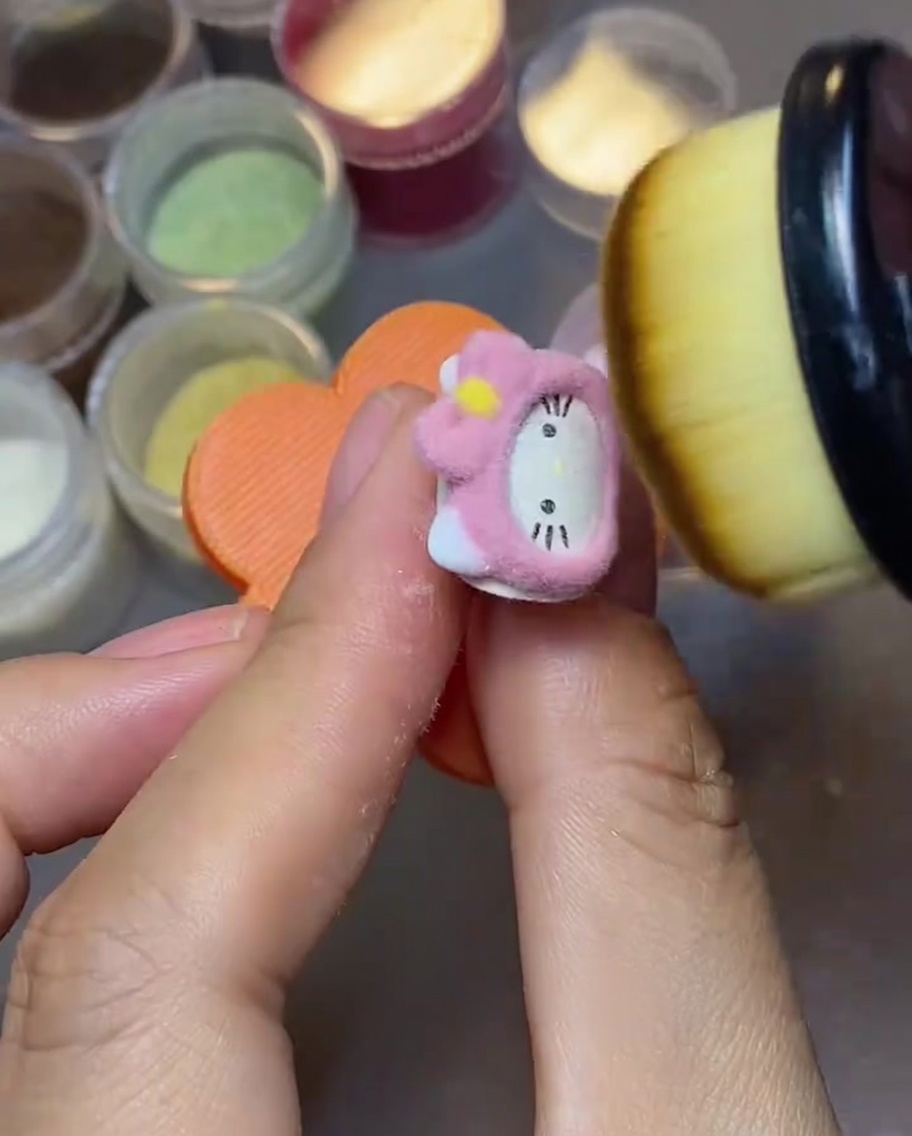 A close-up of a fluffy makeup brush sweeping away loose pink velvet powder from the surface of a Hello Kitty nail charm.
