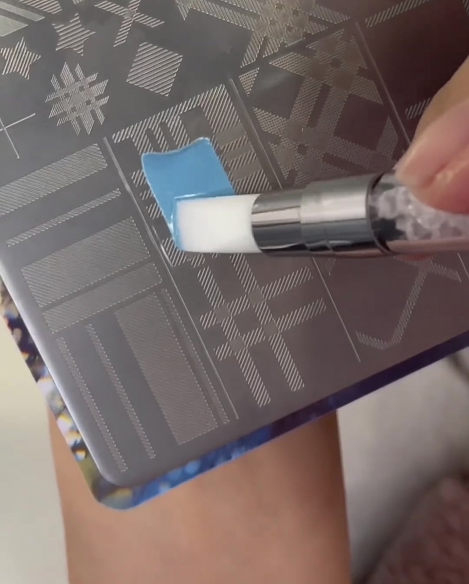 A brush applying light blue polish over a plaid pattern section on a metal nail stamping plate.