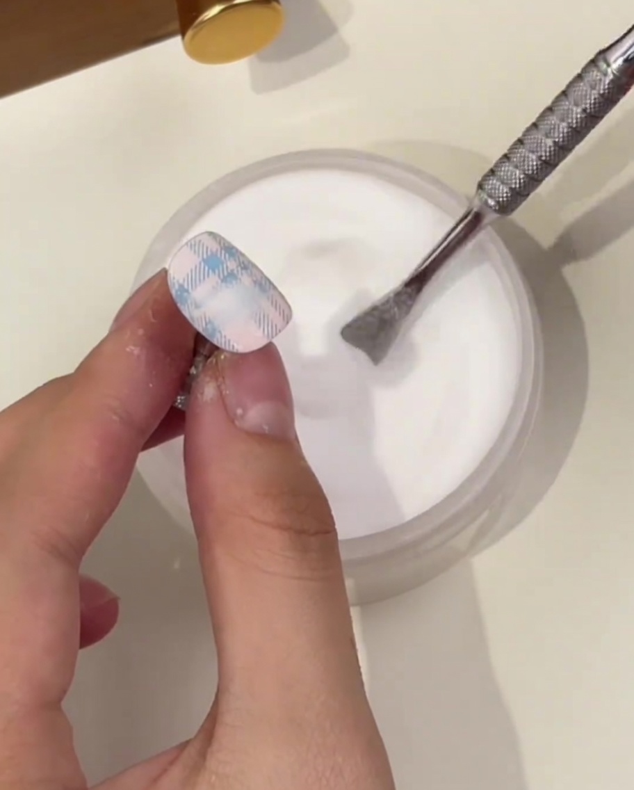 A close-up of a hand holding a nail with a blue plaid pattern, using a metal cuticle pusher to sprinkle clear crystal powder over the wet design above a jar.