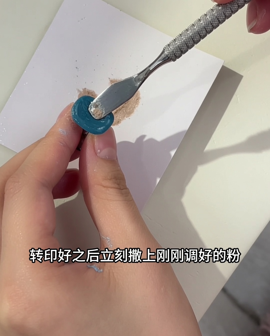 A metal tool applying a second layer of mixed glitter powder over a turquoise nail with a floral design.