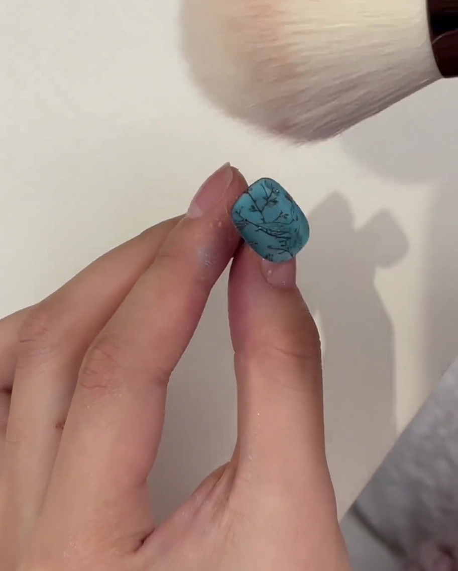 A fluffy makeup-style brush dusting off excess powder from a turquoise nail after curing.