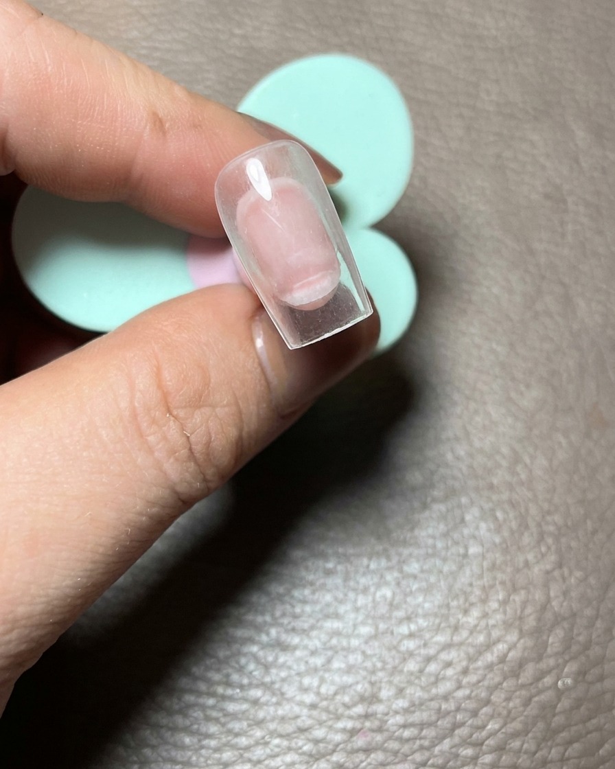 A close-up view of a clear nail tip held by a hand, showing the initial placement of three pink gel dots.