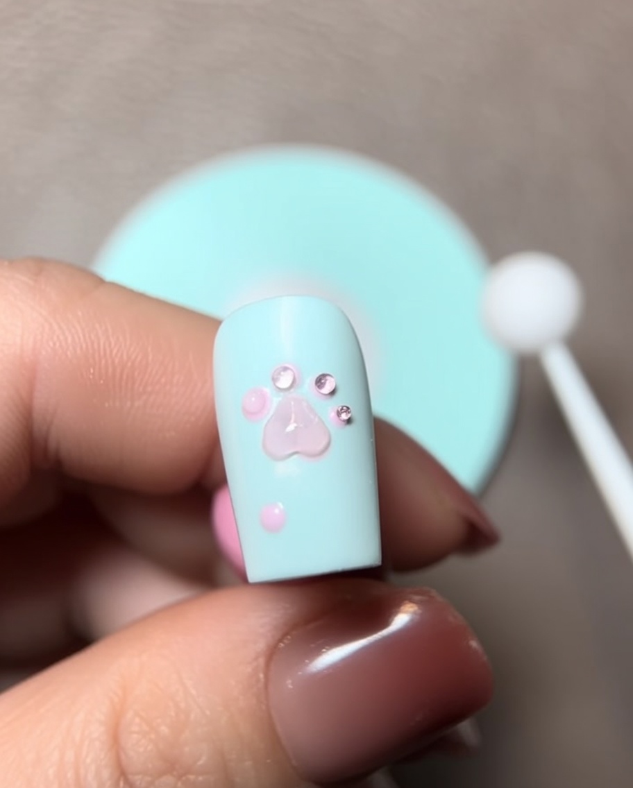 A close-up view of a brush adding a bead of sculpting gel to the 3D paw design on a nail.