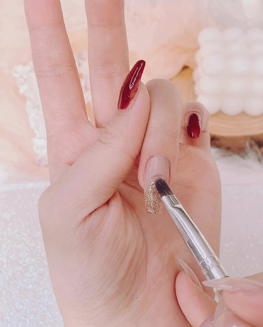 A nail brush applying clear, thick rhinestone glue over a gold glitter Christmas tree shape on the fingernail.
