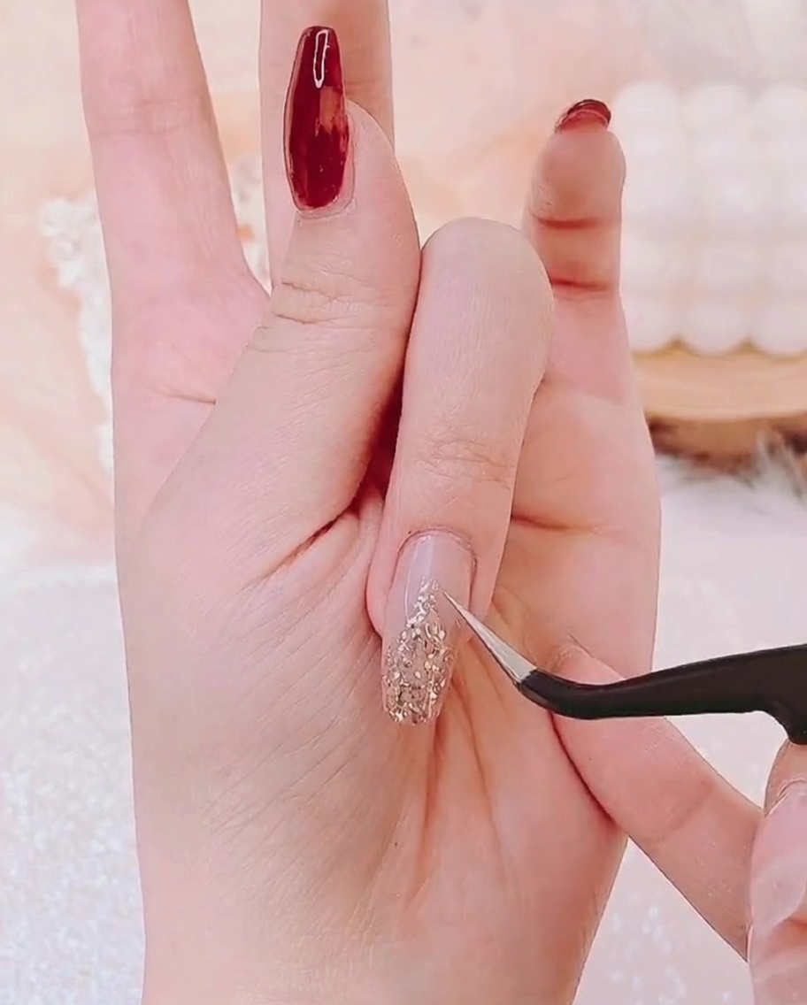 Tweezers placing a small gold star charm at the tip of the gold glitter base on a fingernail.