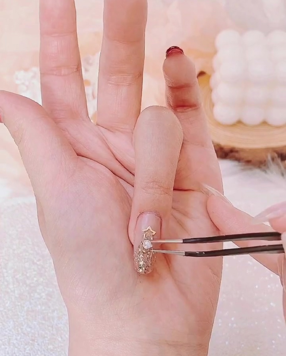 Tweezers positioning a tiny pearl below a gold star charm among other small rhinestones on a fingernail.