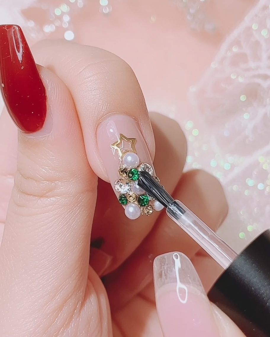 A gel brush applying clear glue around the edges of a 3D rhinestone and pearl Christmas tree design on a nail.