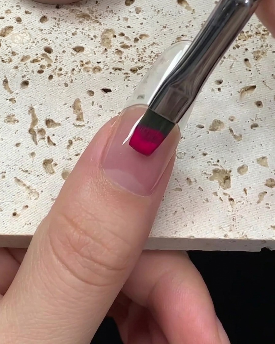 A close-up view of a brush applying vibrant magenta gel polish to a nail, starting from the cuticle.