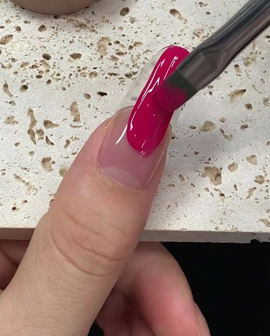 The brush smoothing out the magenta gel polish to ensure full, even coverage on the nail.