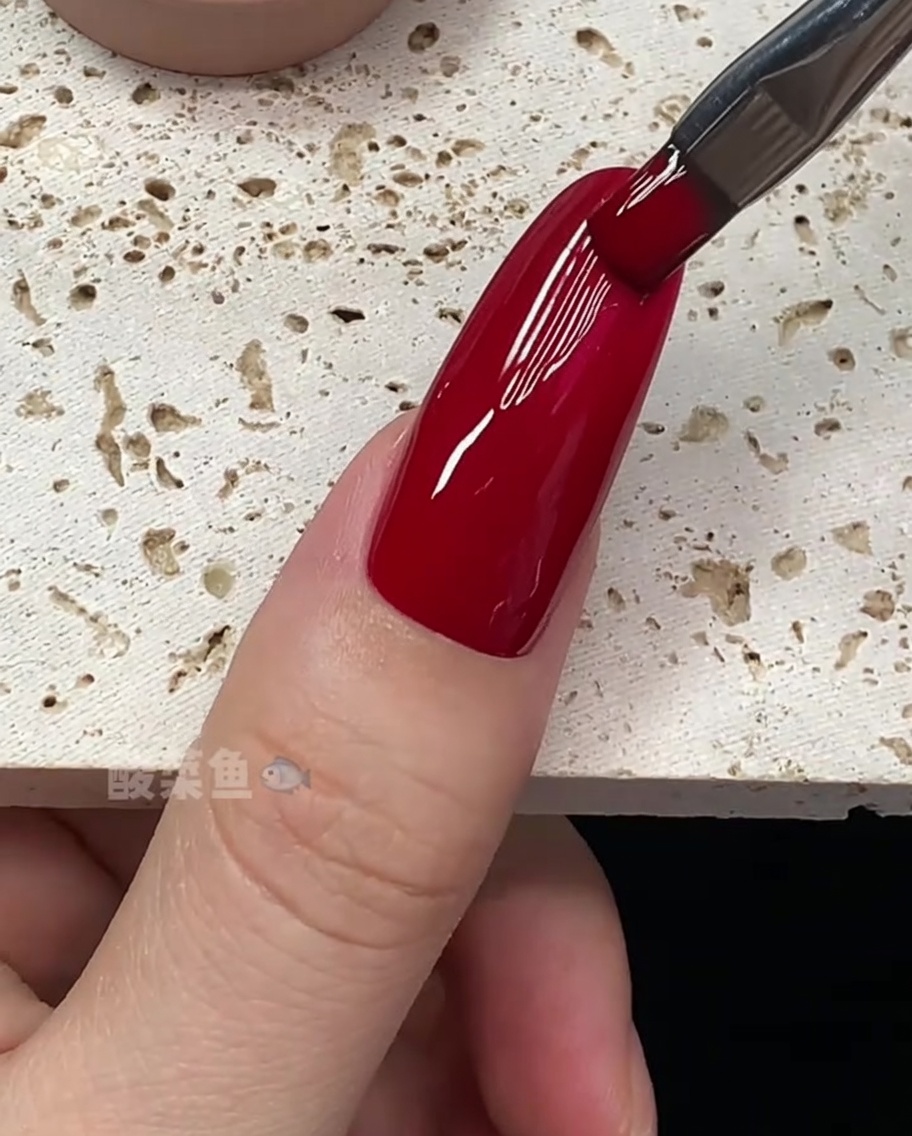 A close-up of a brush perfecting the edges of a nail coated in deep red gel polish.