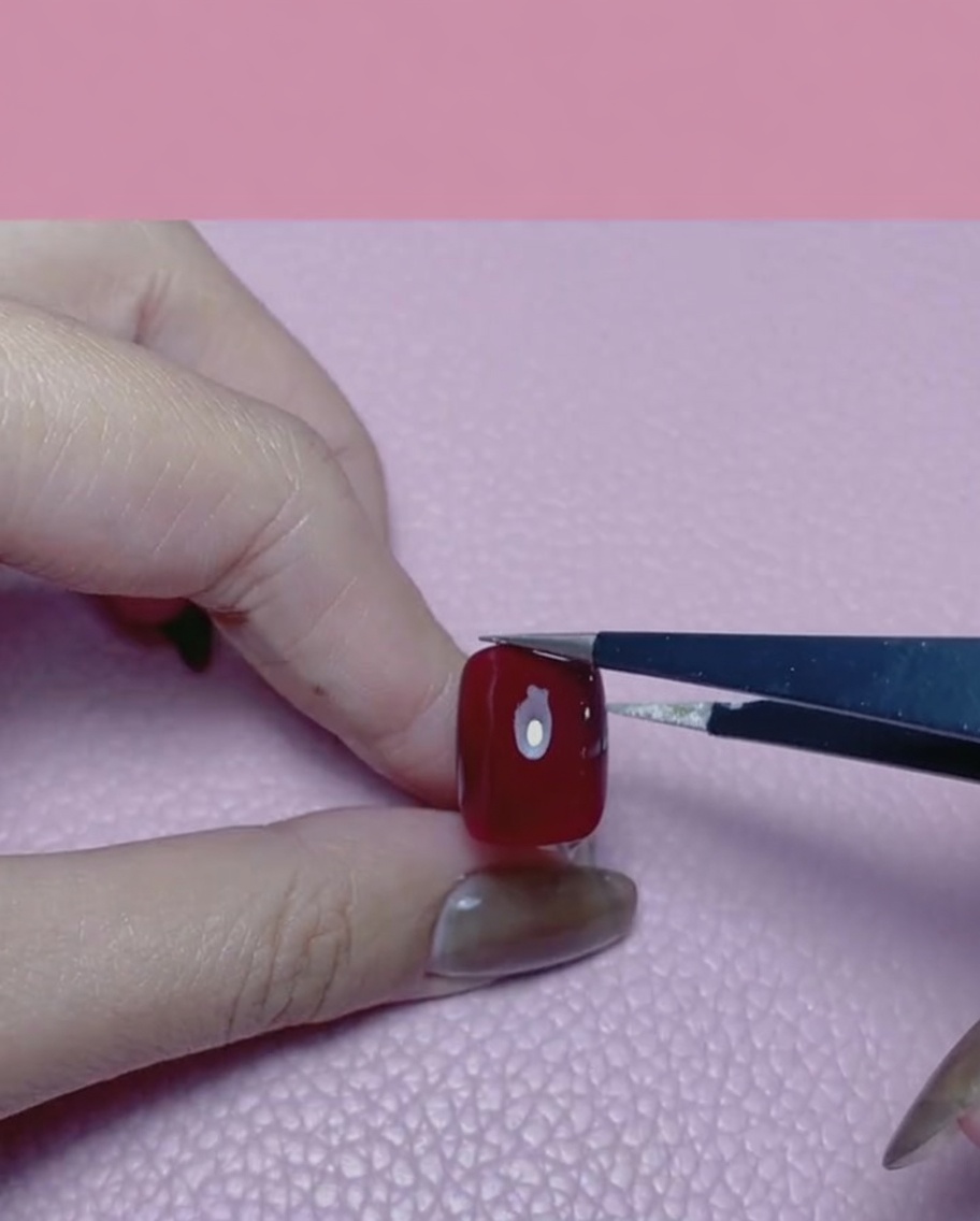 A hand using tweezers to apply a piece of gold foil onto a glossy red fingernail.