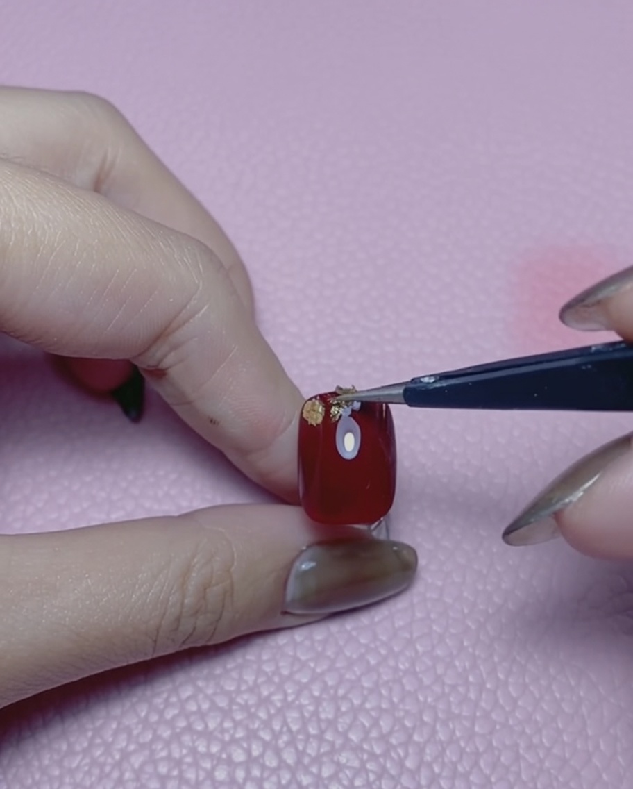 A pair of tweezers holding a small piece of gold foil over a red painted nail.