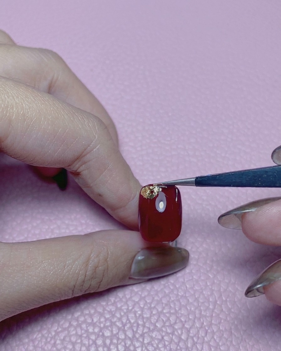 Tweezers placing a third piece of gold foil on a bright red nail.