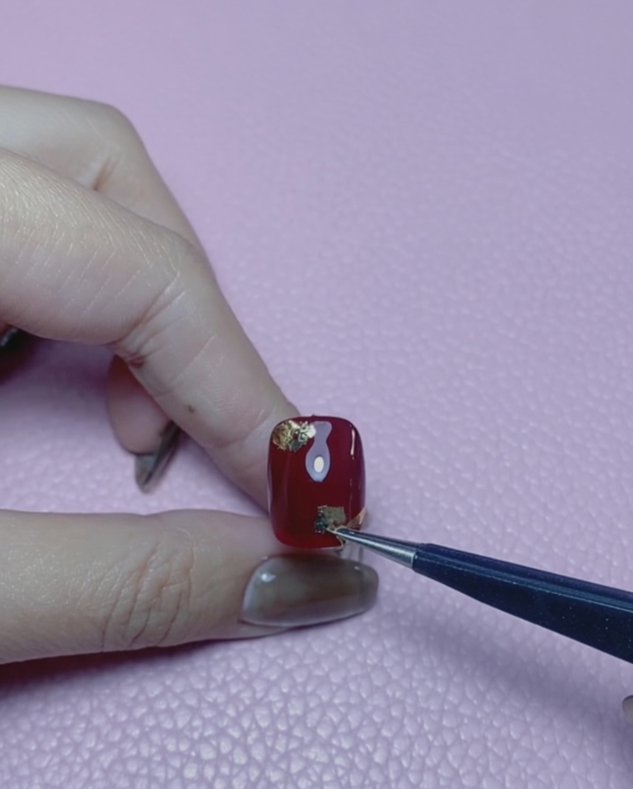 A silicone-tipped nail tool pressing gold foil flakes flat onto a red nail.
