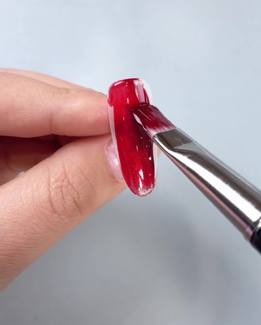 A brush applying red cat-eye gel to the center of a clear nail tip.