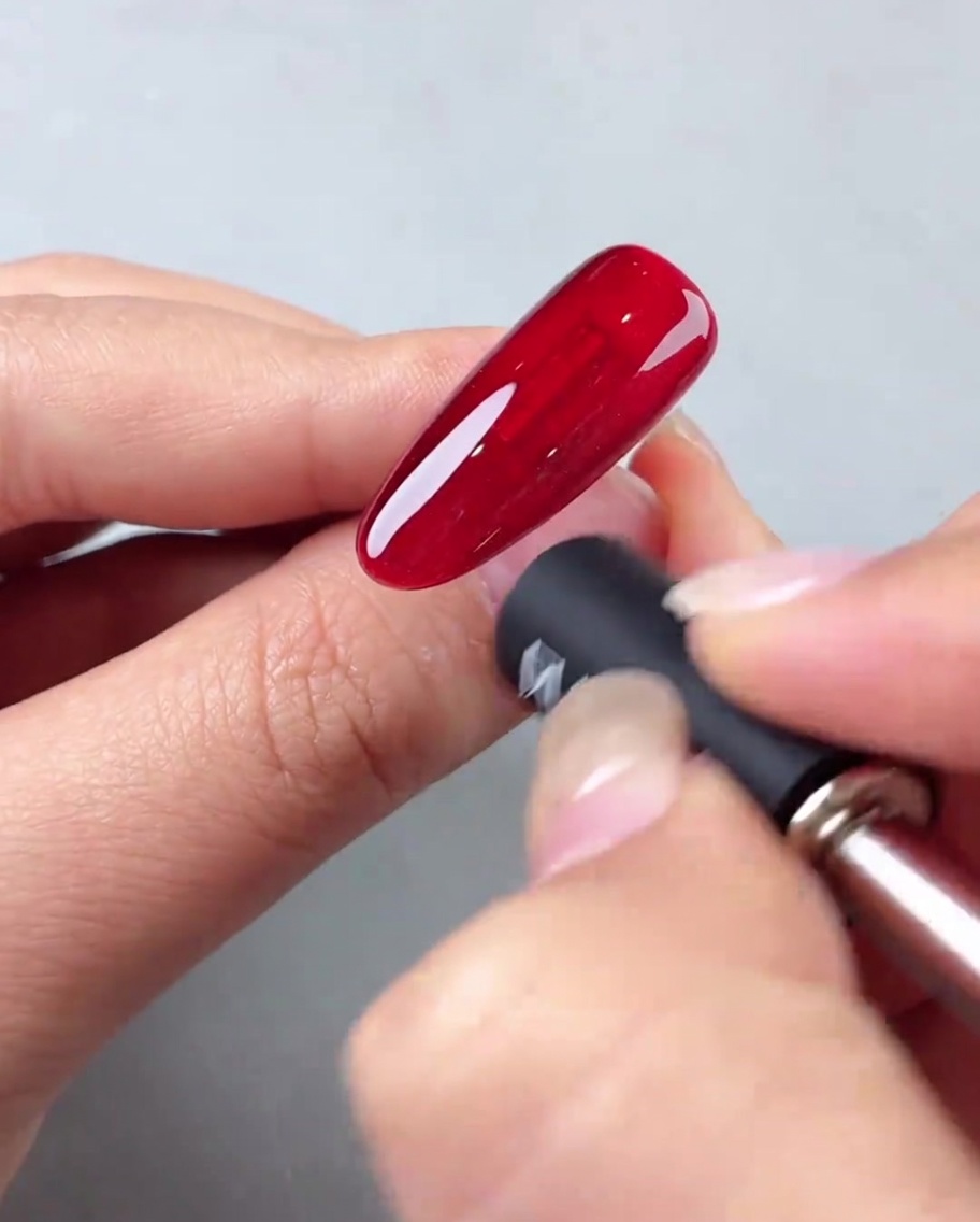 A nail magnet being held at an angle against a red cat-eye gel nail to create a diagonal light effect.
