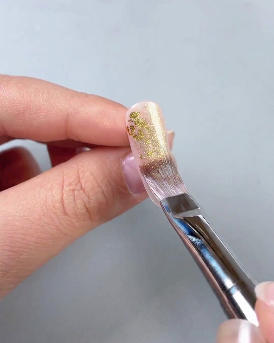A close-up view of a brush applying shimmering gold gel over gold foil pieces on a nail surface.