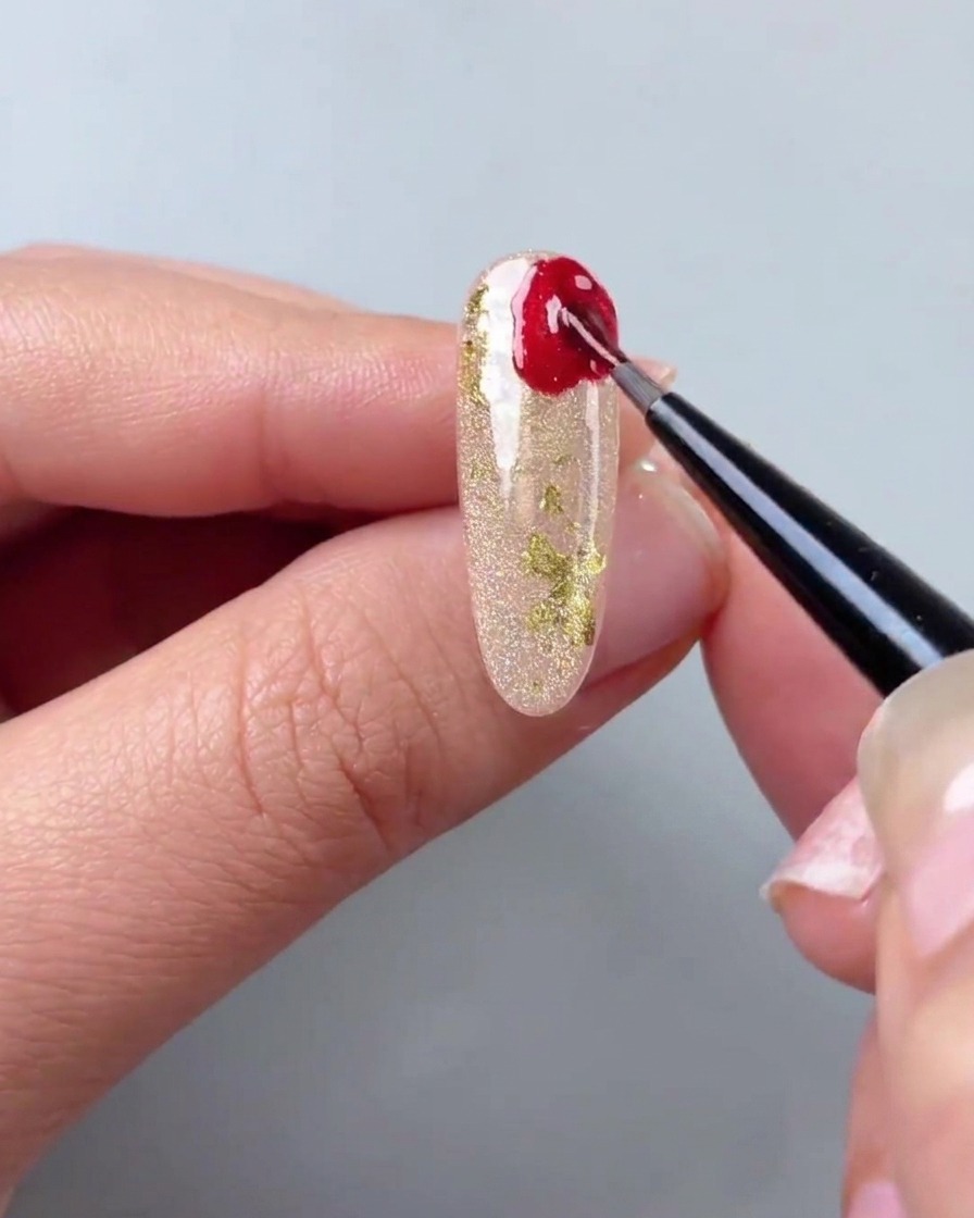 A fine detail brush applying red cat-eye gel in irregular shapes to the corner of a shimmering gold nail.