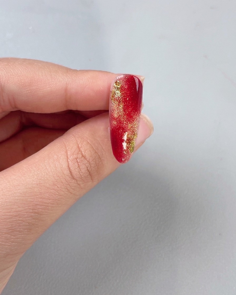A brush applying a clear top coat over a red and gold gradient nail.