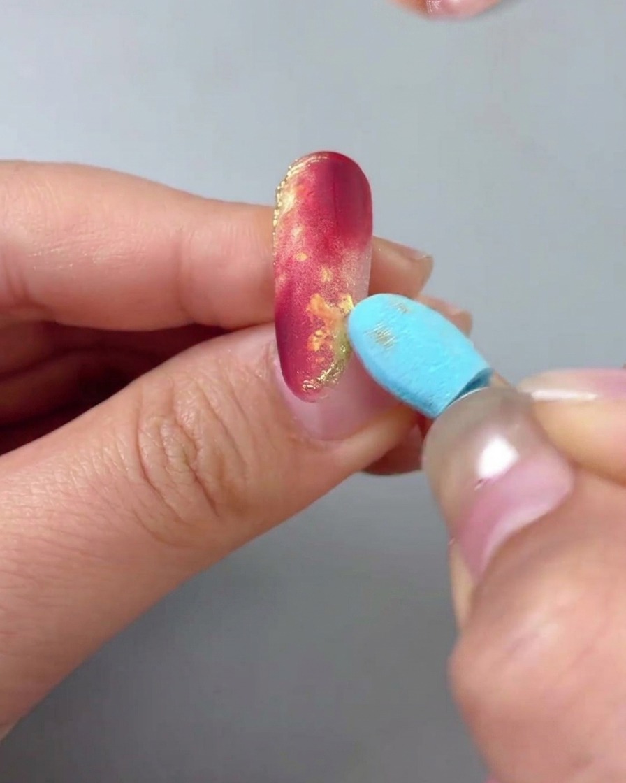 A close-up of a hand with a red and gold marbled nail, while a blue sponge tool gently buffs the surface to remove excess gold powder.