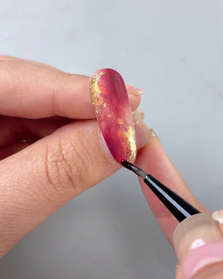 A thin detail brush applying a clear isolating top coat over a beautifully decorated red and gold marble nail.