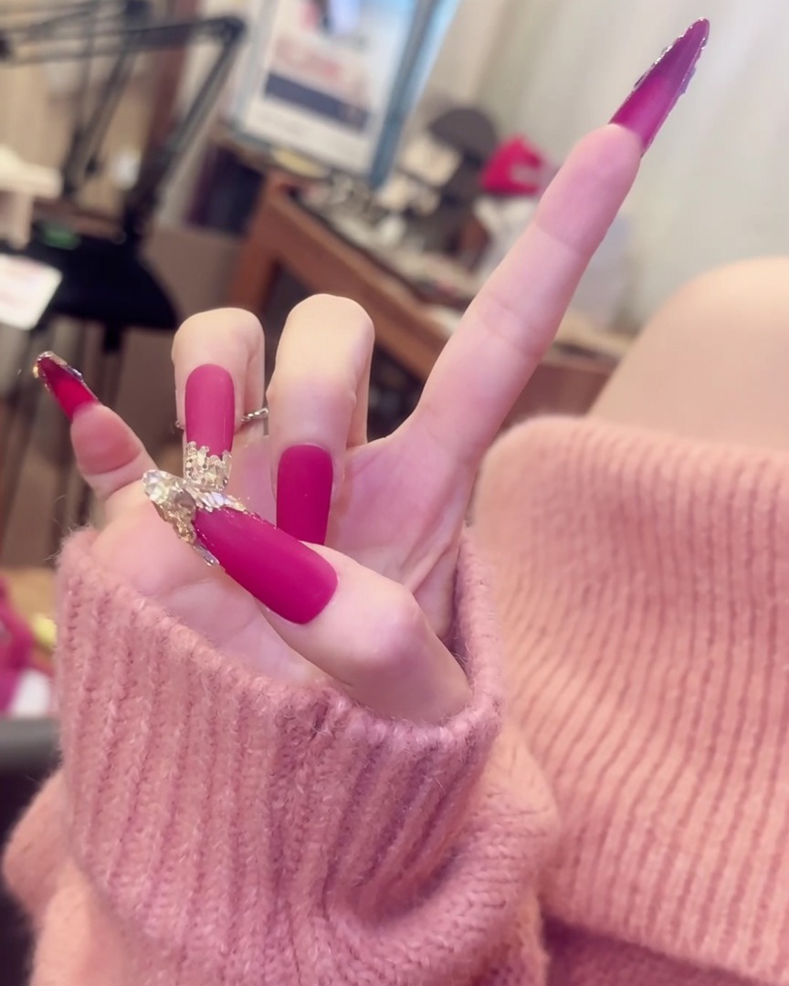 A hand with fingers splayed, showcasing long matte pink nails with crystal accents alongside elegant silver ring accessories.