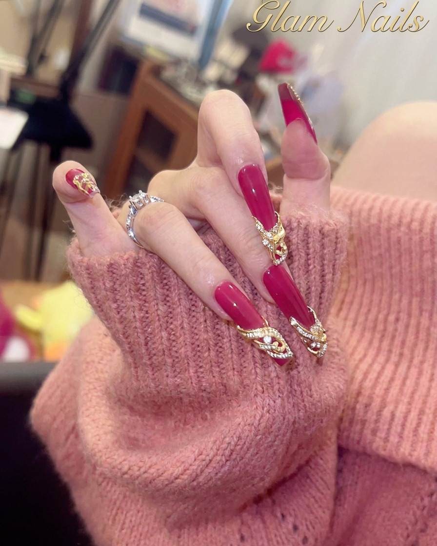 A close-up view of a thumb nail featuring a glossy magenta base and a gold-toned crystal embellishment near the cuticle.