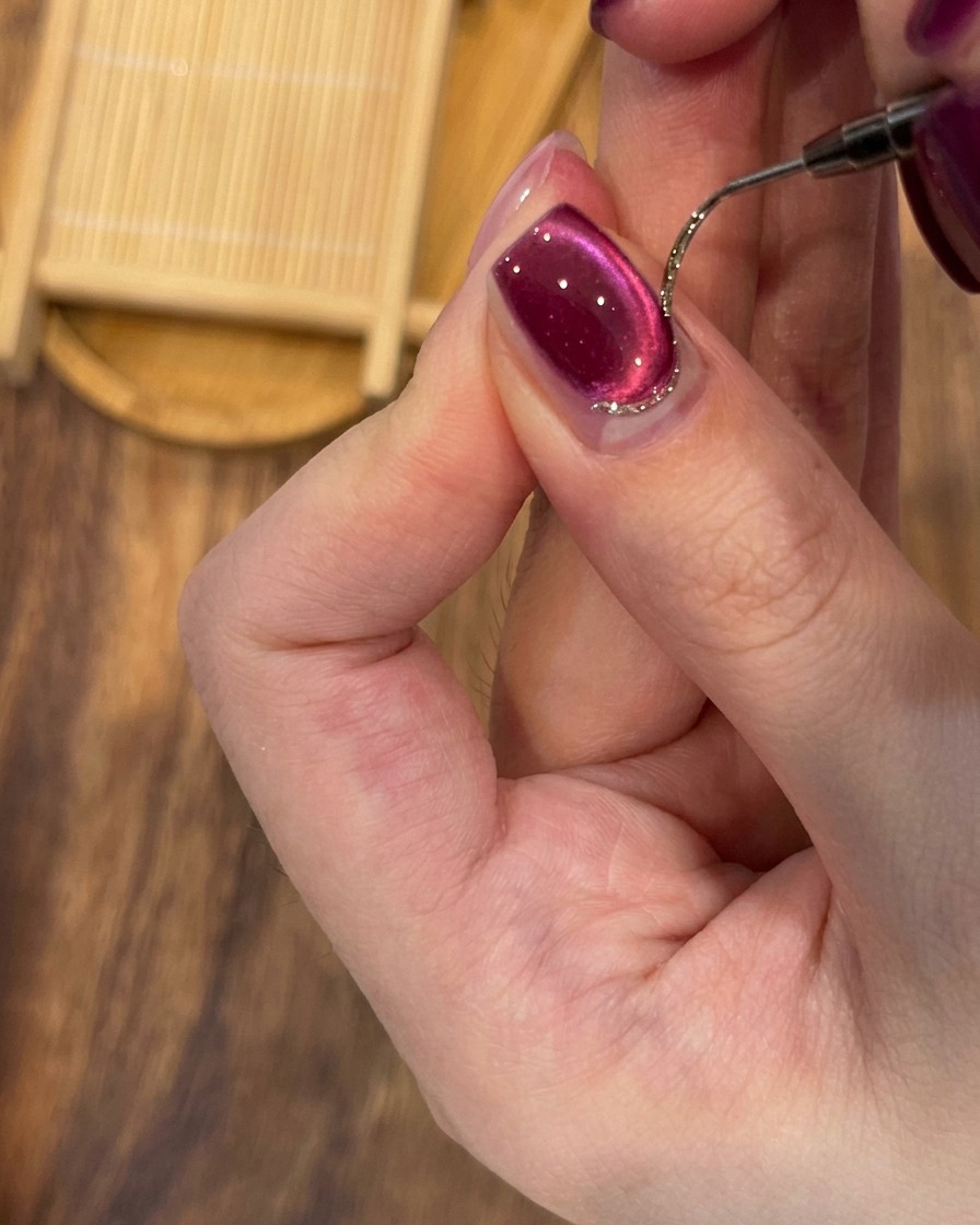A close-up view of a metal cuticle pusher tool being used to gently push back the cuticle on a fingernail.