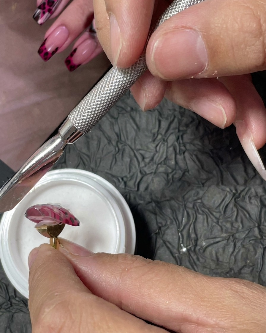 A manicure tool being used to sprinkle fine white acrylic powder over wet red gel dots on a pink nail.