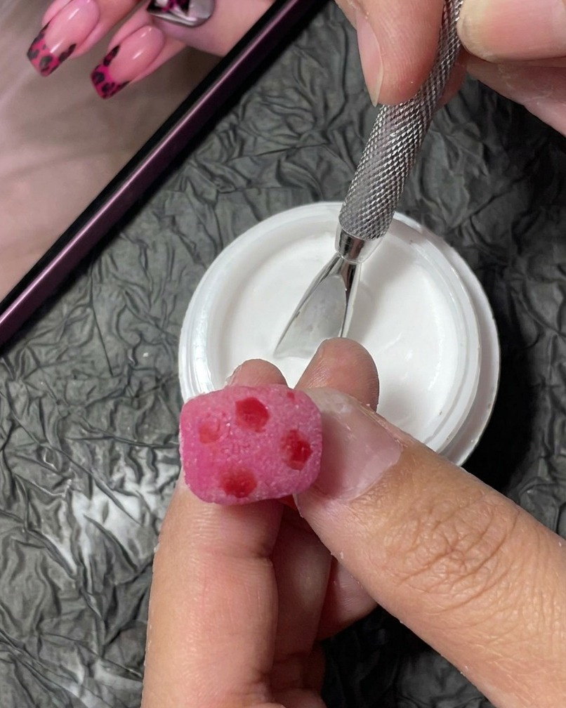 A close-up view of a fine liner brush painting small, irregular black spots onto a pink textured nail.