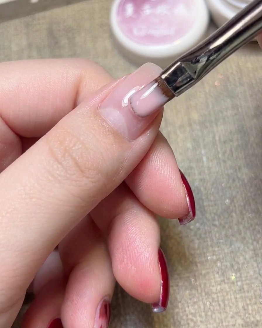 A close-up view of a flat gel brush applying light pink jelly polish onto a natural nail.