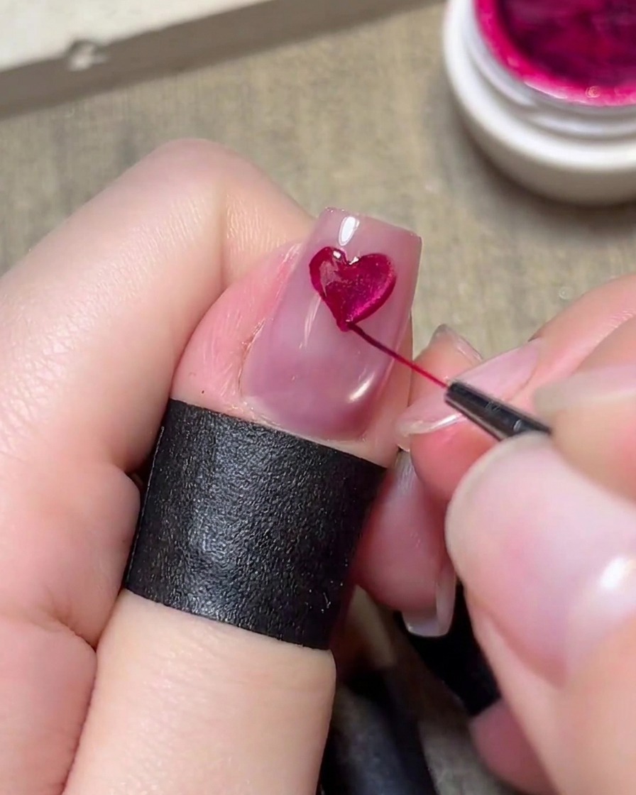 A fine liner brush carefully painting a magenta heart on a light pink nail.