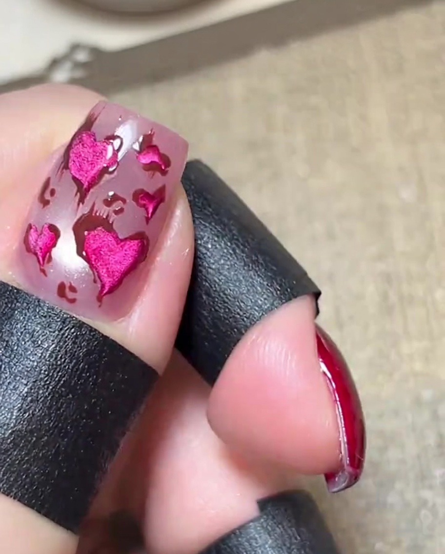 Close-up of a glossy pink nail featuring metallic pink hearts outlined in brown, with delicate, thin brown lines extending outward from the designs.