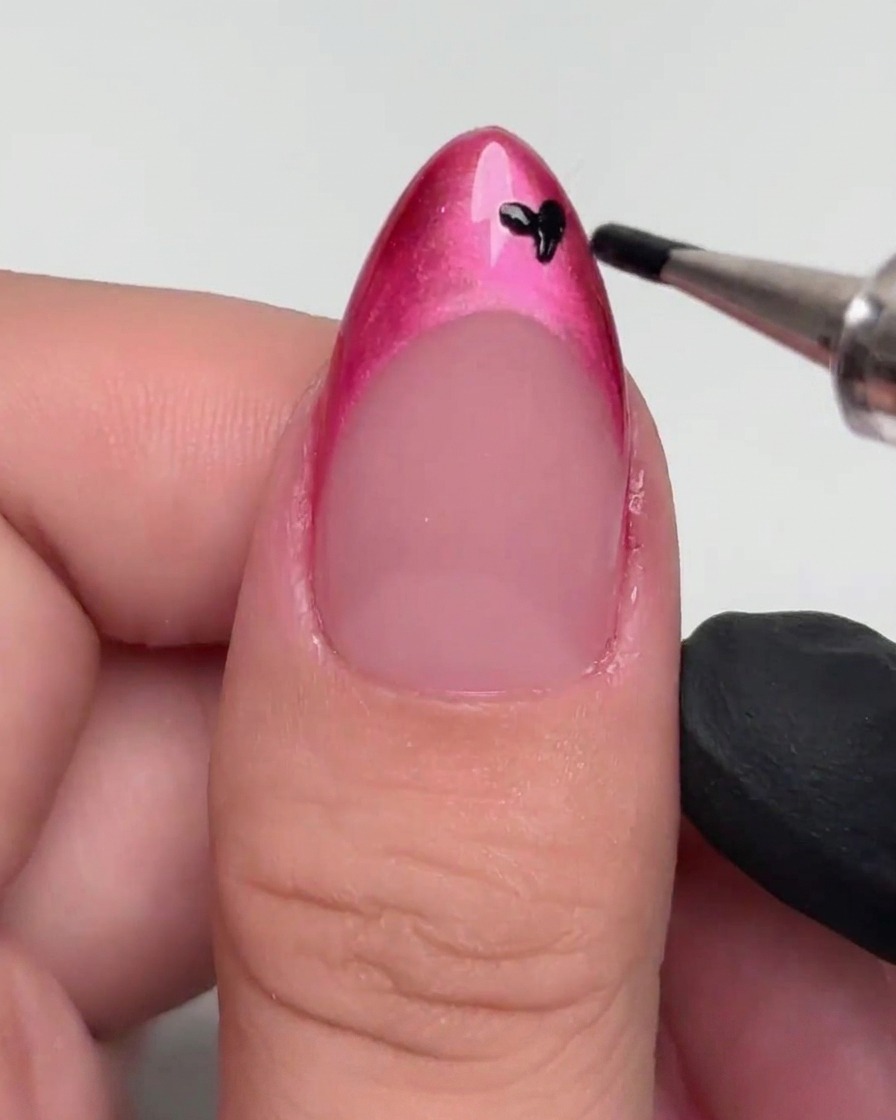A dotting tool applying small black spots onto a magenta-painted French tip.