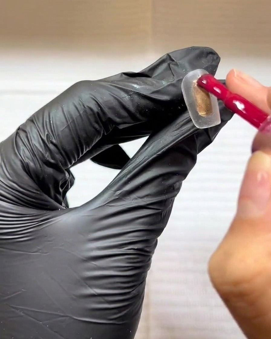 A gloved hand using a gel polish brush to apply a rose red color to a clear nail tip.