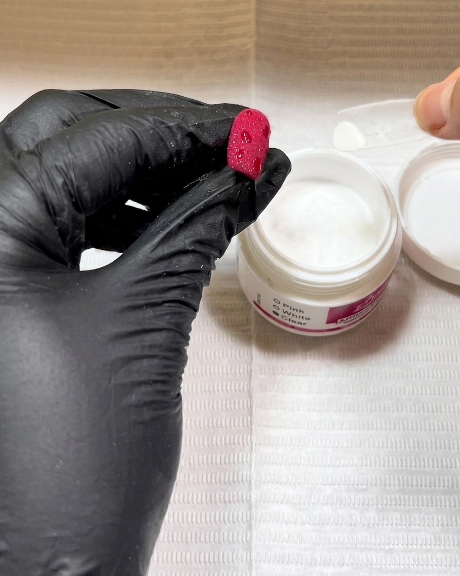 Gloved hands holding a red nail over a jar of clear powder, sprinkling the powder onto wet gel spots.