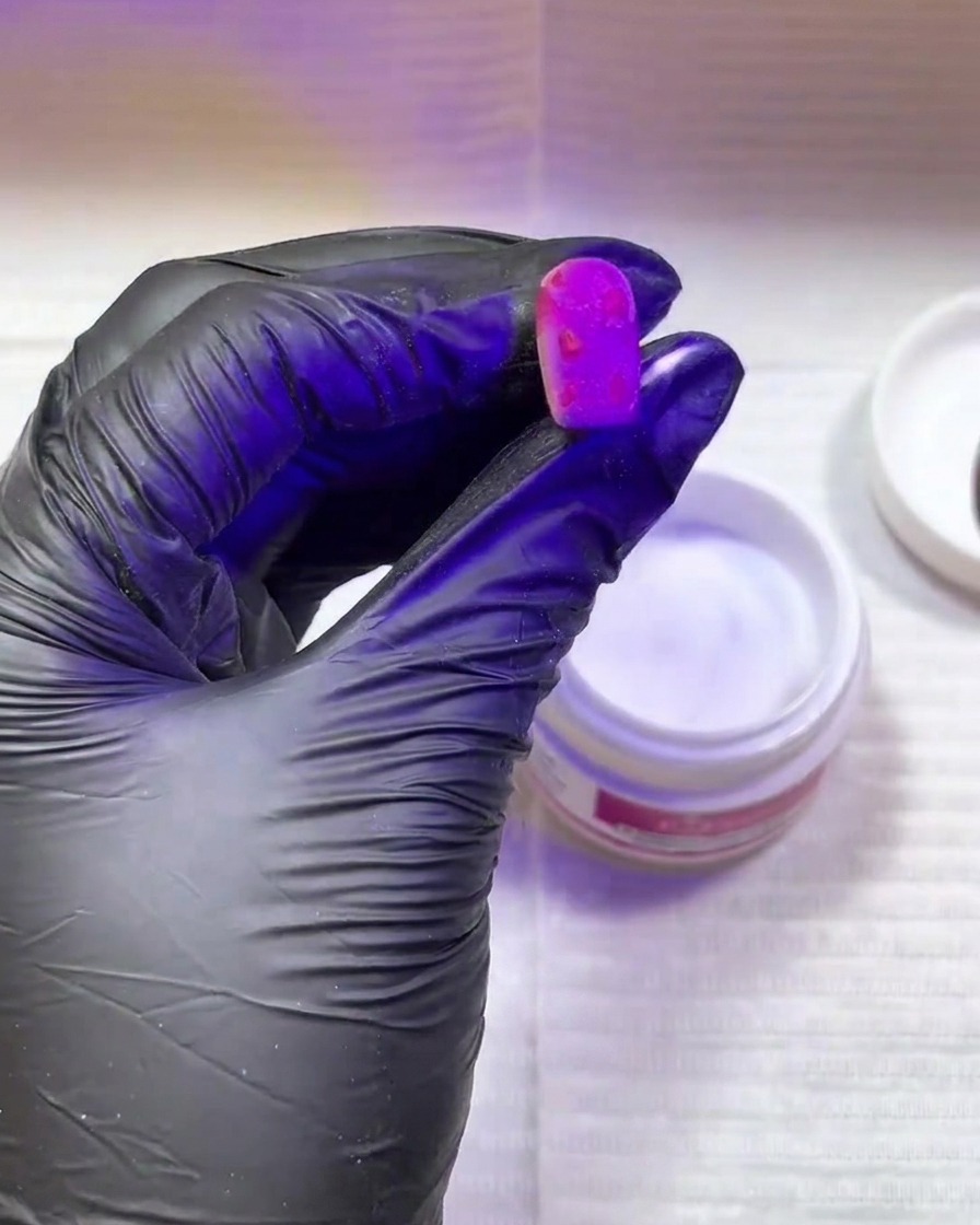 A hand wearing a black glove holding a bright pink nail with textured spots under a purple UV/LED curing lamp.
