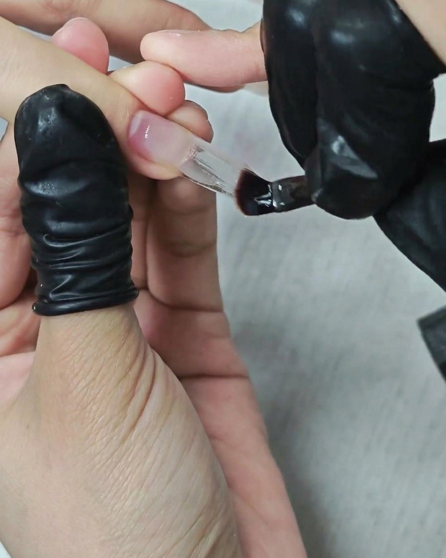 A manicurist using a flat brush to apply and shape thick clear builder gel over a square nail extension.