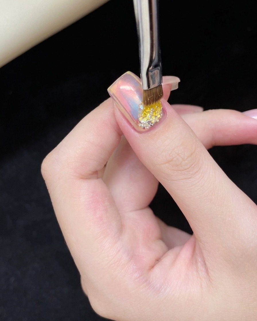 A small nail brush applying a dab of clear adhesive gel onto the bottom portion of a pink cat-eye nail.